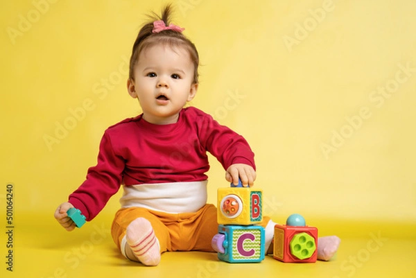 Fototapeta Girl 1 year old, playing with educational cubes with the English alphabet. The girl is sitting on the floor on a yellow background.