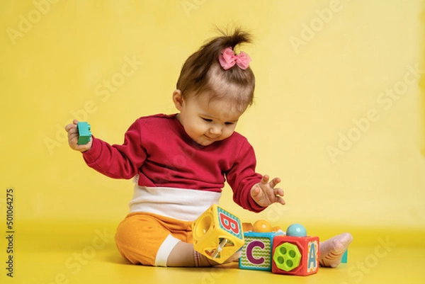 Fototapeta Girl 1 year old, playing with educational cubes with the English alphabet. The girl is sitting on the floor on a yellow background.