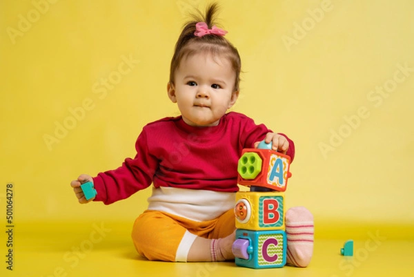 Fototapeta Girl 1 year old, playing with educational cubes with the English alphabet. The girl is sitting on the floor on a yellow background.