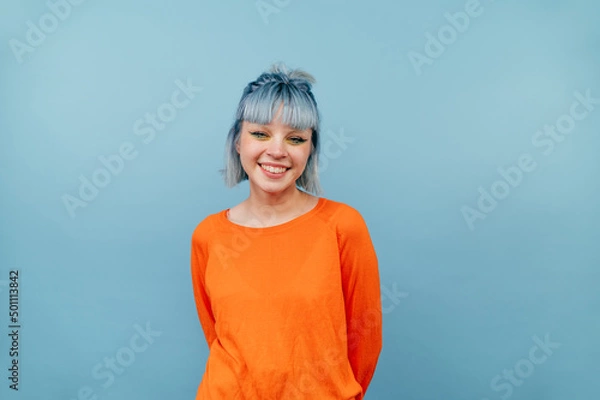 Fototapeta Smiling hipster girl with colored hair stands on a blue background with a happy face and looks at the camera.