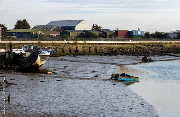 Obraz A small working boat is pushed into the River Medway in Rochester, on a sunny October morning in 2021.  A number of other boats are on the muddy foreshore, with industrial buildings in the background.