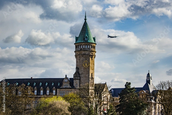 Obraz A Plane Coming in for a Landing at the Luxembourg Airport Flies Past the Spuerkeess Bank Clock Tower in Luxembourg City, Luxembourg