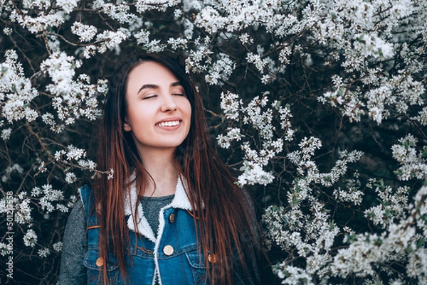 Fototapeta Beautiful girl stands with closed eyes and smiling on a background of flowering spring trees. The girl inhales the scent of spring flowers. Copy paste background