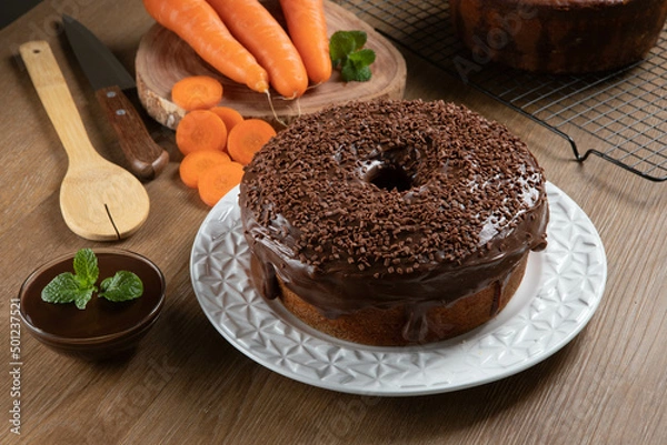 Fototapeta Brazilian carrot cake with chocolate frosting on wooden table with carrots in the background