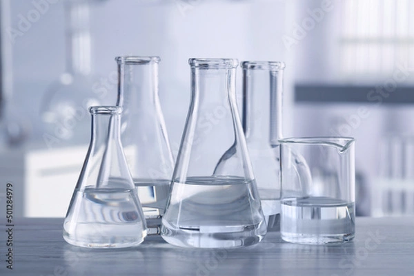 Fototapeta Different laboratory glassware with transparent liquid on wooden table against blurred background