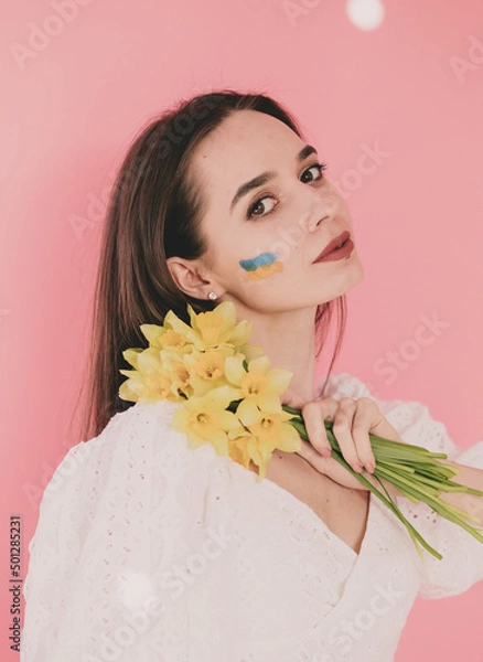 Fototapeta Woman of model appearance with the flag of Ukraine on her face holds in her hands yellow flowers on a pink background