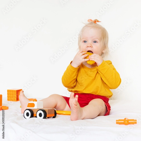 Fototapeta A little Caucasian girl is playing with toys on a white background. 1 year old, the child is wearing an orange T-shirt and playing with orange toys. High quality photo
