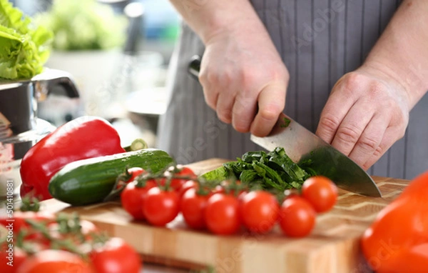 Fototapeta Man chef cutting greenery on wooden board, add ripe vegetable to salad