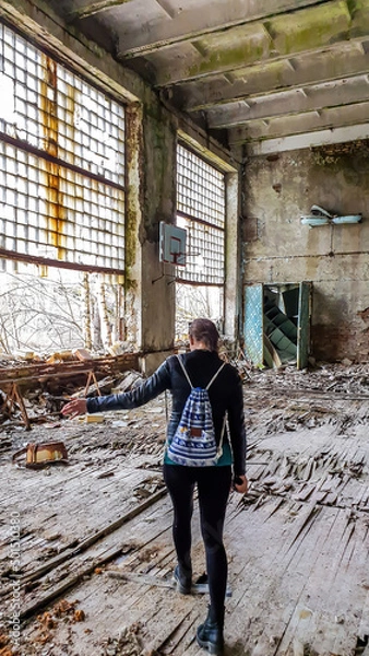 Fototapeta Girl in black outfit walks around the rotten floor in an abandoned sports hall in Pripyat, Ukraine, after the Chernobyl explosion. The floor is completely rotten. Lots of decaying object on the floor