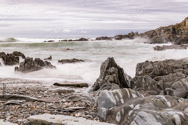 Obraz A rocky and pebbly beach in North Devon, with the sea crashing onto the beach.  A big wave is approaching in the distance.