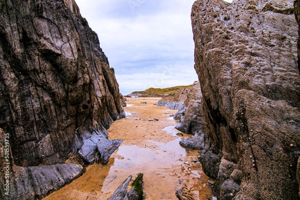 Obraz A sandy channel, revealed by low tide, leads through the rocks towards the sea and headland in the distance.