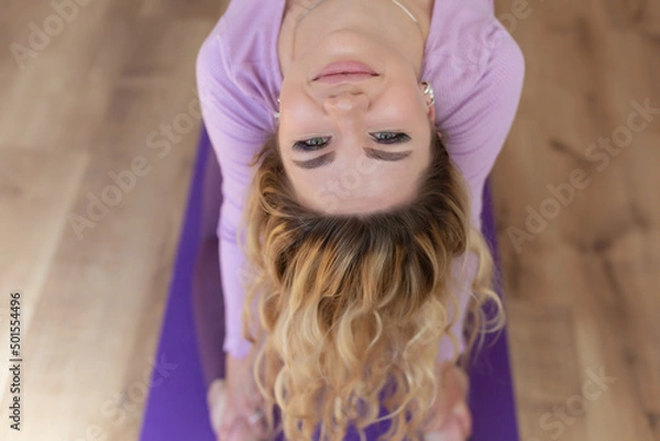 Fototapeta A young blonde girl performs sports exercises in gymnastics, yoga or Pilates at home or in the studio on a mat. Top view of a woman's face with a smile. Close up portrait. Healthy lifestyle.