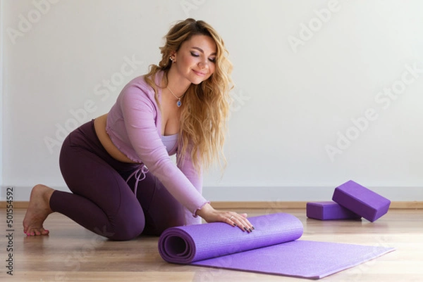 Obraz Young woman rolling lilac mat after gymnastics, yoga or Pilates sports exercises at home or in the studio. A side view of a slender, positive blonde girl on floor near a window on white background.