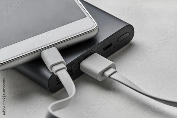 Obraz The smartphone is charging from external powerbank with cable. White background, closeup, selective focus