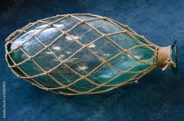 Fototapeta On  blue background is  large glass bottle covered with  woven net of coarse rope.