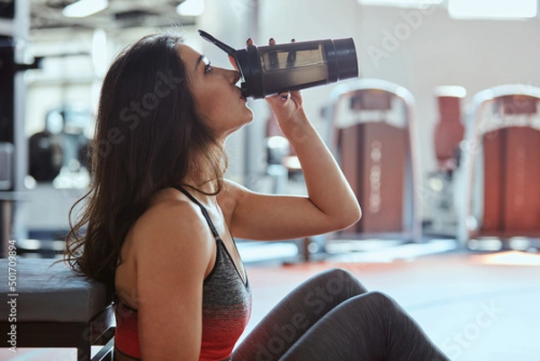 Obraz Young active woman taking a break in the gym and drinking protein shake.