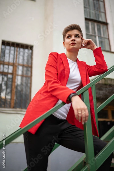 Fototapeta A stylish model girl with short brown hair in a red jacket and a white T-shirt, posing for a photographer standing on a wooden staircase and leaning on her elbows with her face propped on her hand