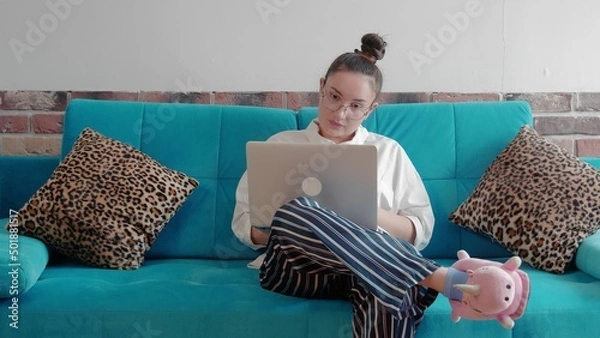 Obraz A young freelance woman in a shirt and slippers, working remotely in a home office, uses a laptop while sitting on the couch. A modern woman communicates in an online conference using a computer.