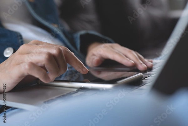 Fototapeta Young casual woman sitting on sofa using mobile phone and laptop computer at home, close up