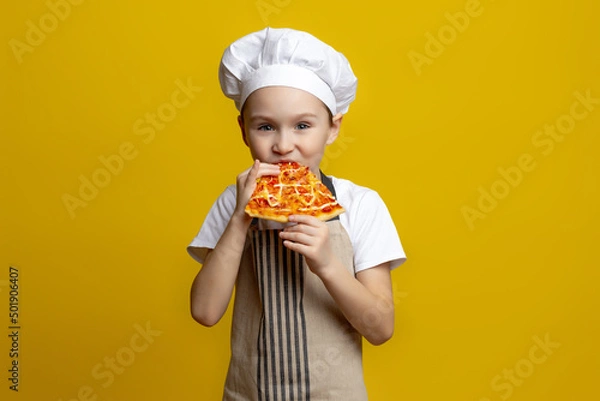 Fototapeta A little funny boy cook of 6-7 years old in a hat and apron holds a rolling pin in his hand for dough and points it aside on a yellow studio background in a black circle. Concept