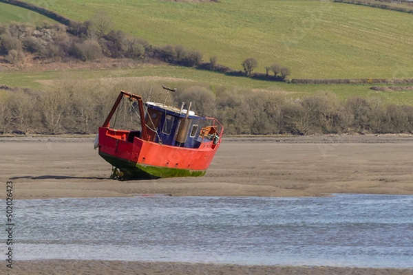 Fototapeta A rusting fishing trawler lays stranded on a sandbank in a tidal estuary. River Teifi, with hilly farmland background. Rippling blue water. Landscape image with space for copy. Poppit Sands, Wales.