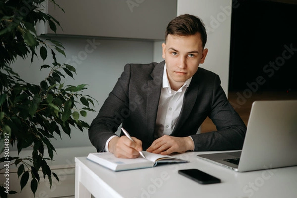 Fototapeta Serious young Caucasian man sitting at desk making notes in notebook