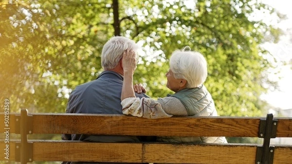 Fototapeta Outdoor photo of senior caucasian heterosexual couple sitting on a bench in the park enjoying beautiful weather. Gray-haired lady in warm vest touches her husband's hair. High quality photo