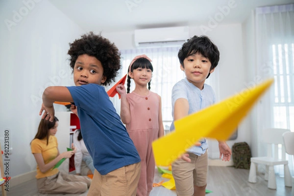 Obraz Asia and African kid playing paper rocket in the kindergarten school classroom. Rocket fly in front of camera.  Learning, enjoy, education, playing concept. Feeling happing and smile during class