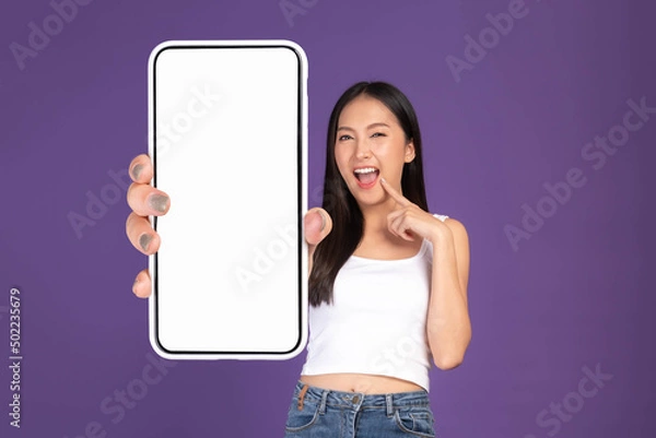 Fototapeta Beautiful Asian brunette woman cute girl in white tank top ,Excited surprised girl showing big smart phone with blank screen , white screen isolated purple background , Mock Up Image