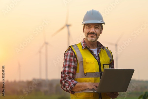 Fototapeta Portrait of power engineer wearing safety jacket and hardhat with laptop computer working at outdoor field site that have wind turbine at the background.