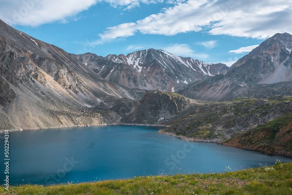 Fototapeta Deep mountain lake of phantom blue color among sharp rocks and high mountains in changeable weather. Wonderful dramatic view from sunlit grassy hill to deep blue alpine lake and rocks under cloudy sky