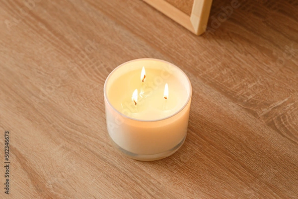 Fototapeta A large scented candle with three burning wicks and melting wax in a glass standing on a wooden table. Side view. Place to copy