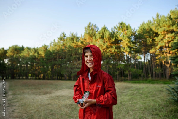 Fototapeta A boy in a red windbreaker against the backdrop of a forest with a compass in his hand.