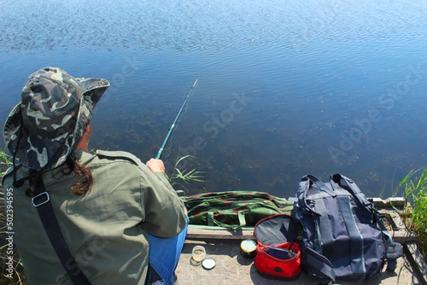 Fototapeta A fisherman with a fishing rod sits on the river bank, rear view on a summer day. Catching fish from the lake on a sunny day