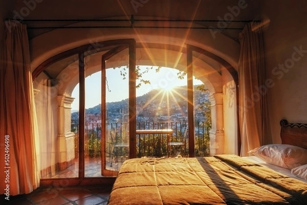 Obraz Outside the windows of the hotel is a city on the slopes of the mountains. The rays of the evening sun and the roofs of houses. Balcony with chairs, glass doors, beds and room interior with colonial a