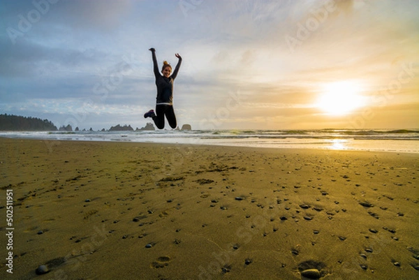 Fototapeta Adventurous athletic woman jumping in the air and smiling at the camera on a Pacific Northwest beach during a beautiful sunset.