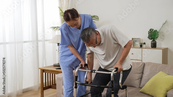 Obraz asia female nursing aide helping injured retired male to use a walker while he is slowly standing up from the couch at home. in-home health care for elderly senior concept