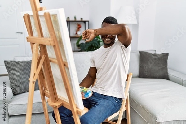 Fototapeta Young african man painting on canvas at home smiling cheerful playing peek a boo with hands showing face. surprised and exited