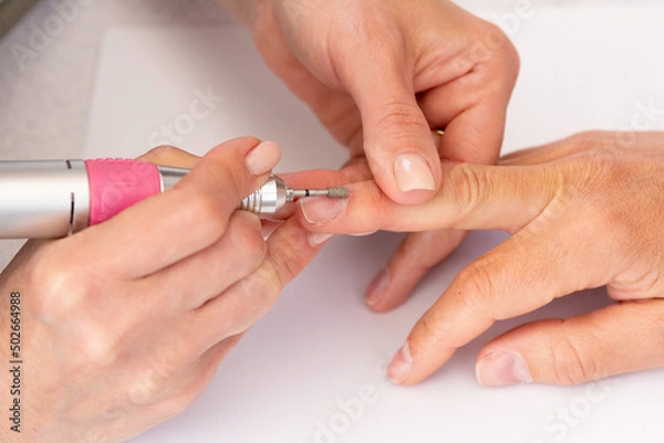 Fototapeta Manicure process in beauty salon, cleaning of nails by a milling cutter. Manicurist filing client's nails at table. Removing the nail plate with a milling machine. nail care concept