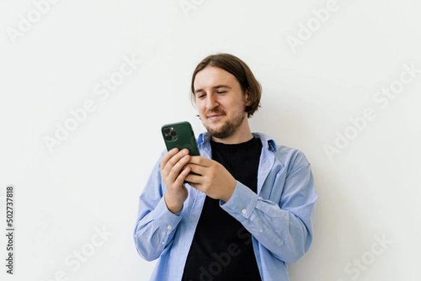 Fototapeta Young smiling businessman watching something on mobile phone isolated on white background. Studio shoot. Copy space