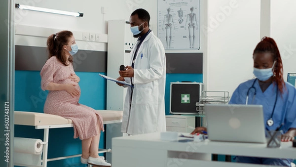 Fototapeta Medical specialist and pregnant woman with face mask at checkup visit talking about bottle of pills. Obstetrician using medicine to help patient with pregnancy belly, giving maternity advice.