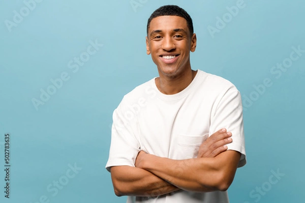 Obraz Smiling young African-american man in white casual t-shirt standing with crossed arms confident and happy, looking at camera with wide toothy smile isolated on blue background