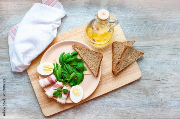 Obraz Plate with spinach leaves, green basil, bacon, eggs and a piece of rye bread. Olive oil. Healthy breakfast. Food for breakfast on wooden background. Flat lay