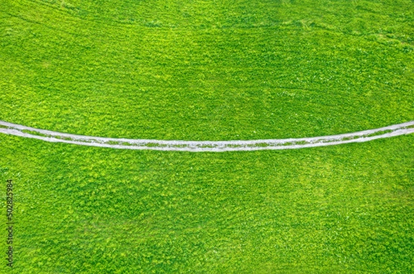 Fototapeta Unpaved countryside road across green field, drone view from above