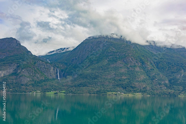 Fototapeta View of the Lustrafjord in Norway.  turquoise sea water, low white clouds above it, cloudy sky, green forest on the mountain slopes, white waterfall 