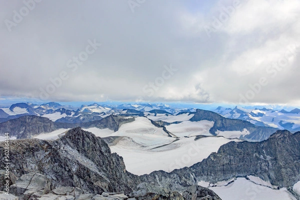 Fototapeta View of the mountain landscape in Jotunheimen national park in Norway from the Galdhopiggen mountain. Grey cloudy sky, grey and brown mountain slopes, white snow and ice of the glaciers