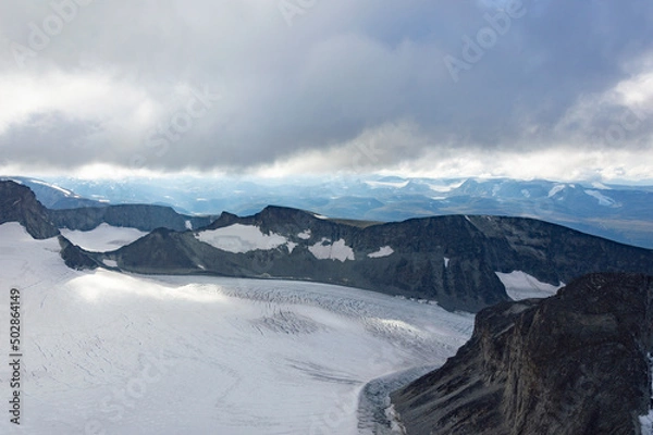 Fototapeta View of the mountain landscape in Jotunheimen national park in Norway from the Galdhopiggen mountain. Blue sky with clouds, grey and biege rocks, mountain slopes, white snow and ice of the glacier