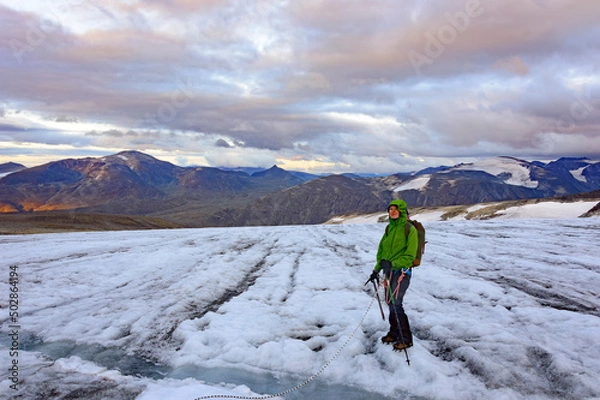 Fototapeta young woman alpinist standing near the crack in the glacier on the way to the Galdhopiggen mountain summit in Norway. White and grey snow and ice, blue sky with clouds and dark mount on the background