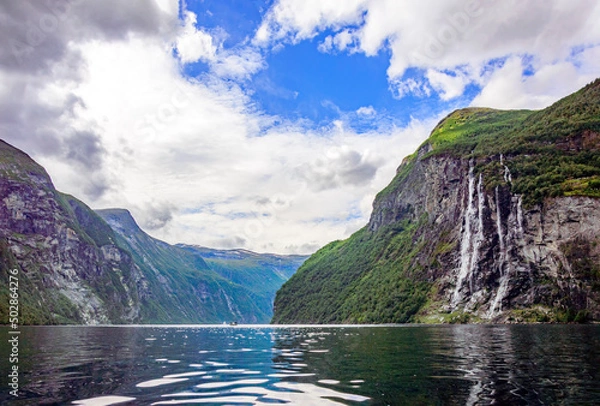 Fototapeta View of Geirangerfjord and the Seven sisters waterfall in Norway from the water.  Blue sky with clouds, green mounain slopes with sunshine and shadows on the forest