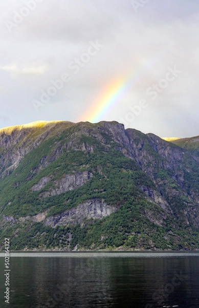 Fototapeta View of Geirangerfjord in Norway. Blue and grey sea water, blue sky with clouds, green and mounain slopes with sunshine and sadows on the forest, rainbow above the summit